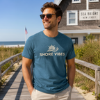 Man wearing a 'Shore Vibes' t-shirt on a beach boardwalk with a building in the background.