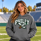 Woman wearing a heather gray Philly pullover hoodie with white and green varsity PHILLY lettering and an eagle carrying a football on the chest