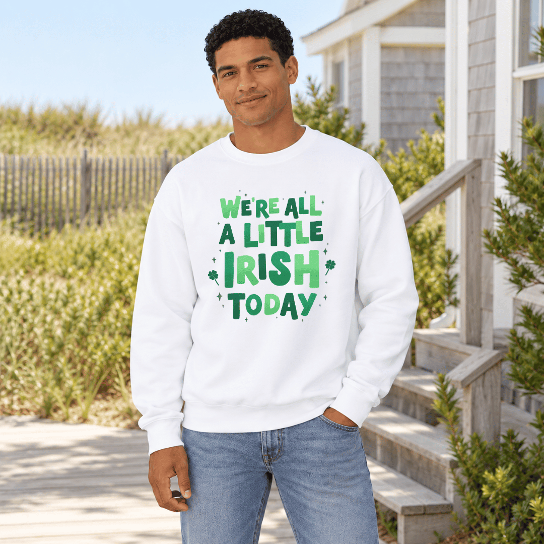 A man wearing a white crewneck sweatshirt with a green 'We're All A Little Irish Today' graphic, standing outdoors in front of a house and dunes.