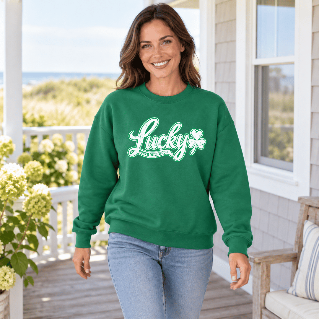 A woman stands on a porch wearing a kelly green crewneck sweatshirt with a white graphic that reads 'Lucky' and 'North Wildwood' alongside a shamrock. She is wearing light blue jeans, and the background shows a coastal home exterior with a window, wooden railing, and ocean view.
