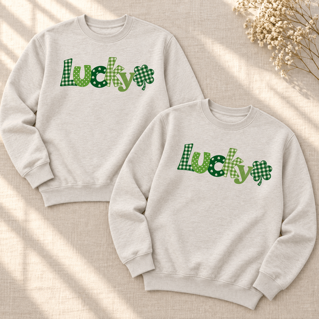 Two Heather Dust crewneck sweatshirts laid flat, showing green Lucky lettering and plaid shamrocks