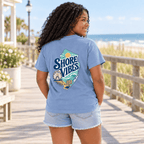 Back view of a girl in a Melon orange Shore Vibes t-shirt on a boardwalk with a ferris wheel in the distance