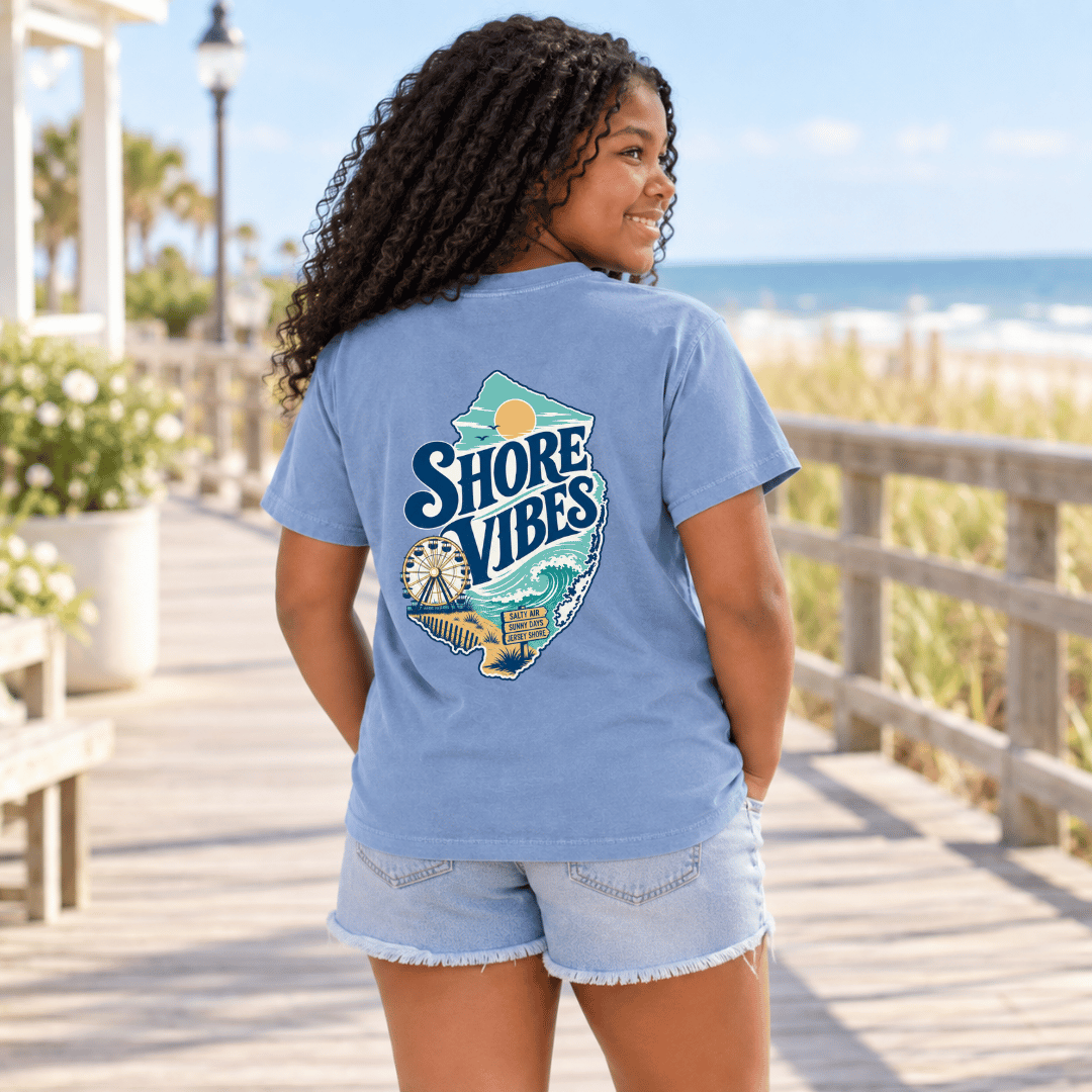Back view of a girl in a Melon orange Shore Vibes t-shirt on a boardwalk with a ferris wheel in the distance