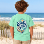 Back view of a boy in an Island Reef green Shore Vibes t-shirt standing on a dune path by the ocean