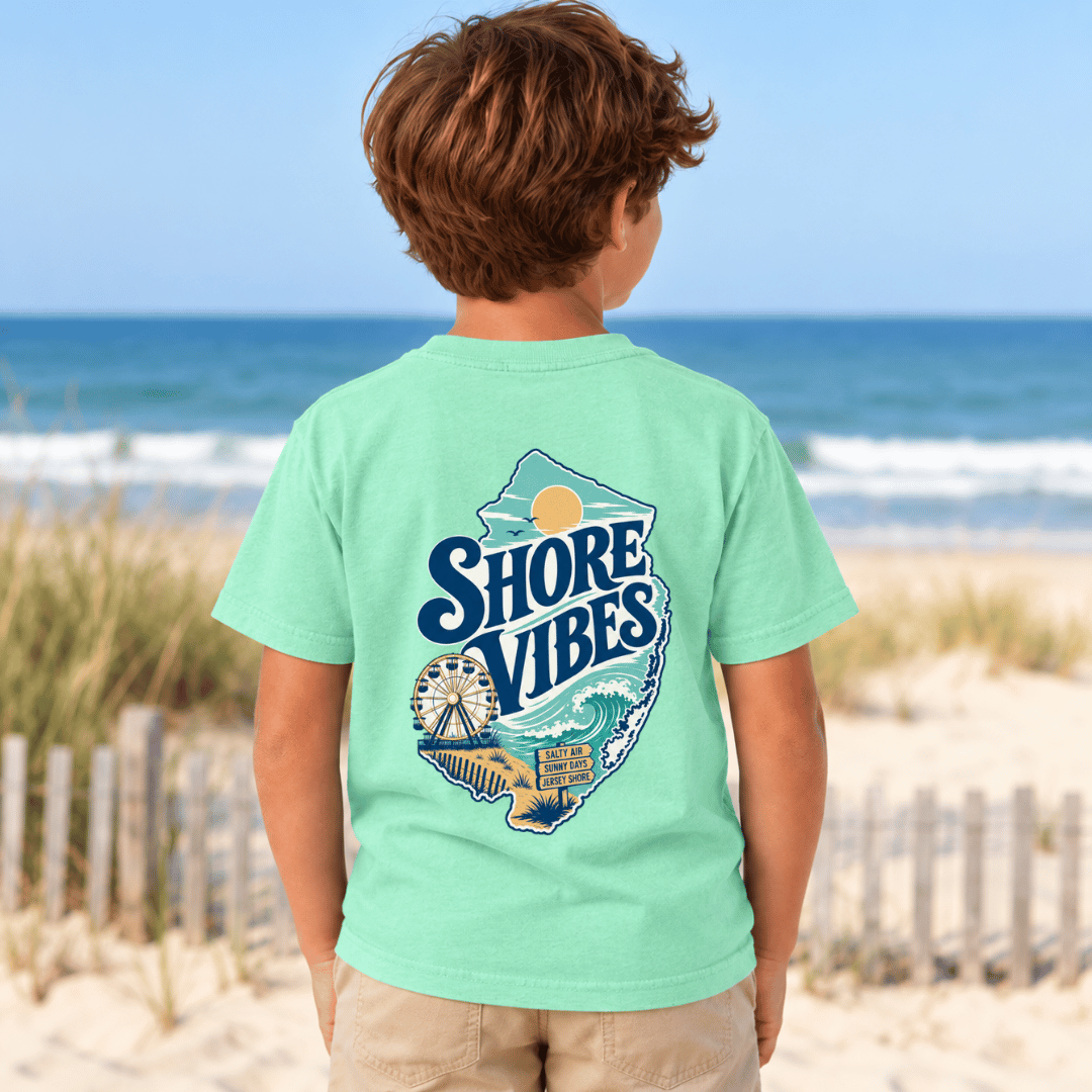 Back view of a boy in an Island Reef green Shore Vibes t-shirt standing on a dune path by the ocean
