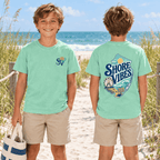 A boy in Island Reef green Shore Vibes t-shirts on a beach path, one showing the front logo and one showing the back graphic