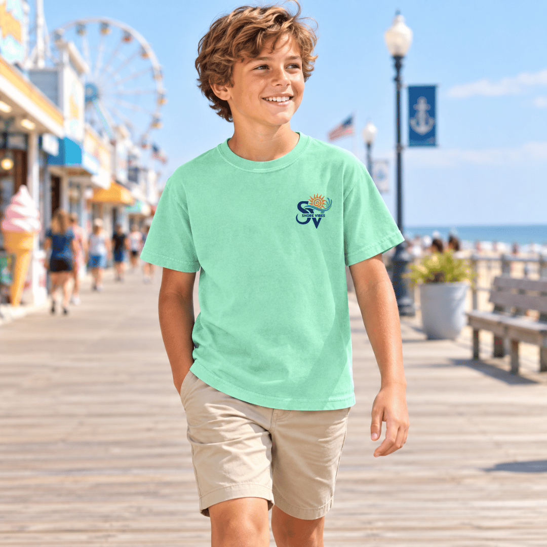 Boy wearing an Island Reef green Shore Vibes t-shirt with the front logo, walking a boardwalk near a beach ferris wheel