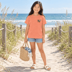 Girl in a Melon Shore Vibes t-shirt carrying a straw bag on a boardwalk path to the beach