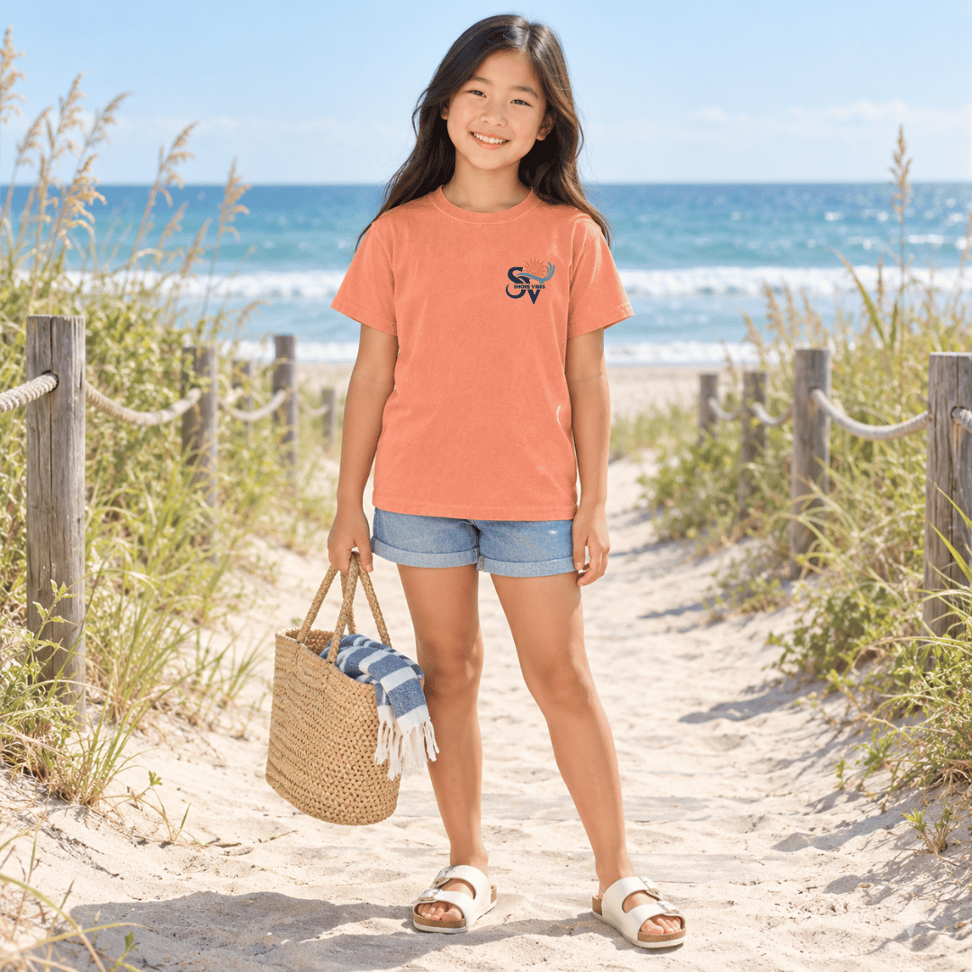 Girl in a Melon Shore Vibes t-shirt carrying a straw bag on a boardwalk path to the beach