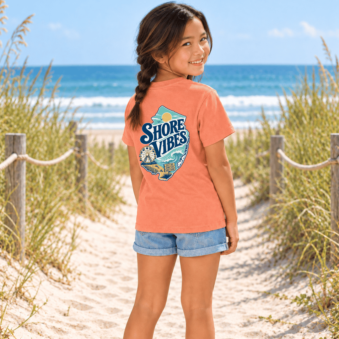 Back view of a girl in a Melon orange Shore Vibes t-shirt walking a boardwalk path toward the beach