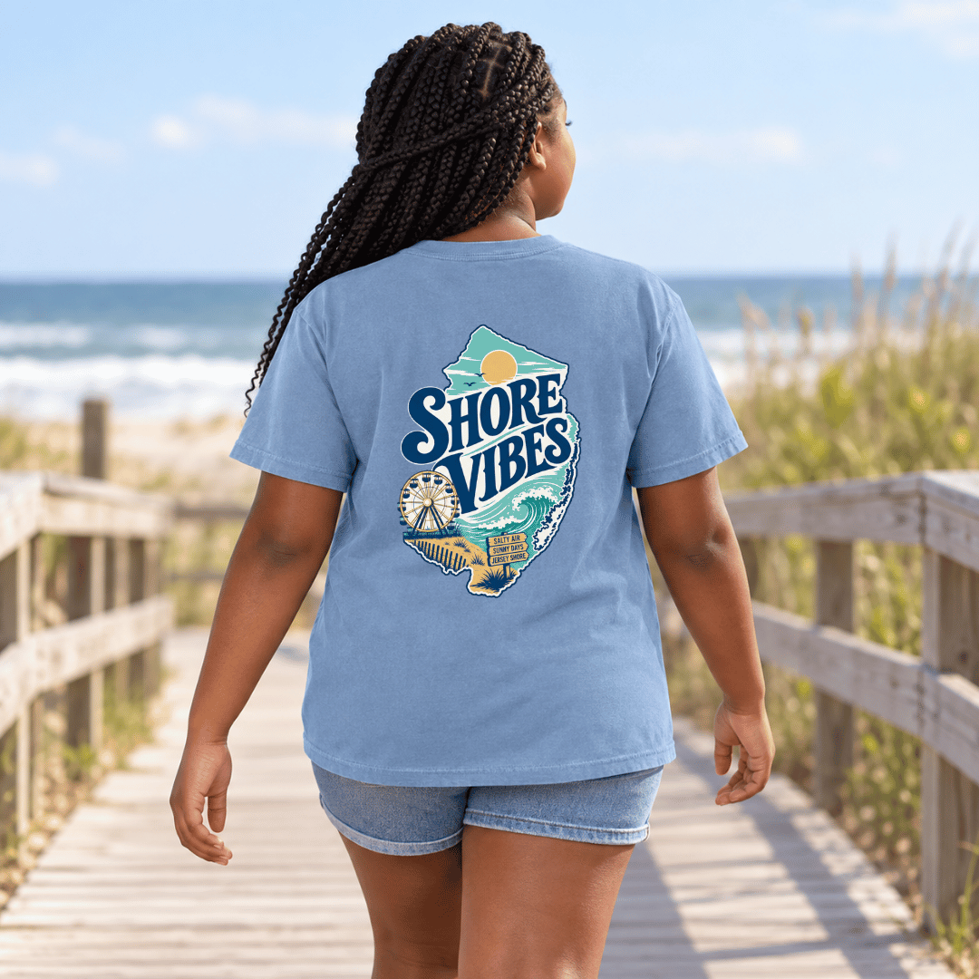Back view of a girl in a Washed Denim blue Shore Vibes t-shirt walking away on a boardwalk near the beach