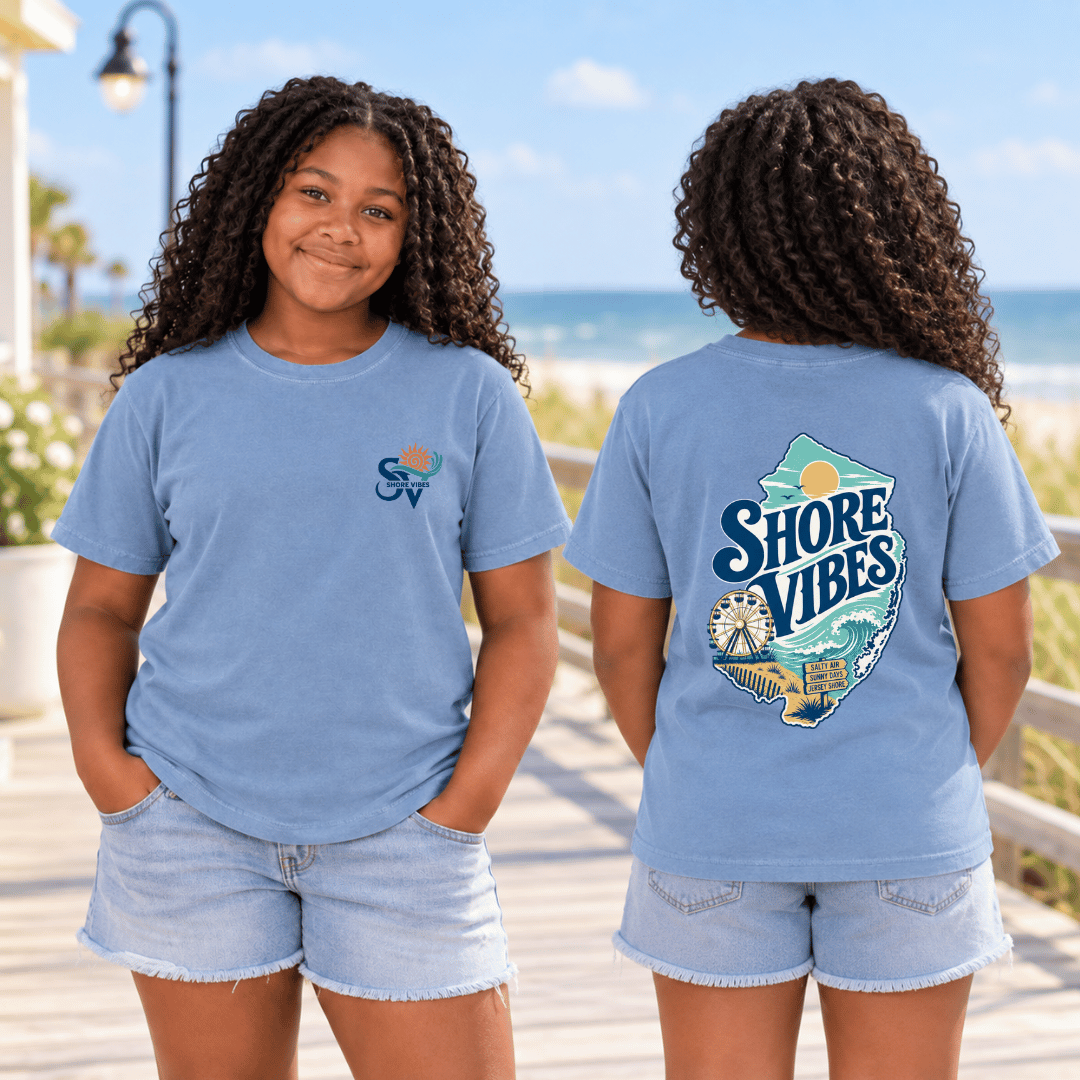 A girl in Washed Denim blue Shore Vibes t-shirts on a boardwalk, one showing the front logo and one showing the back graphic