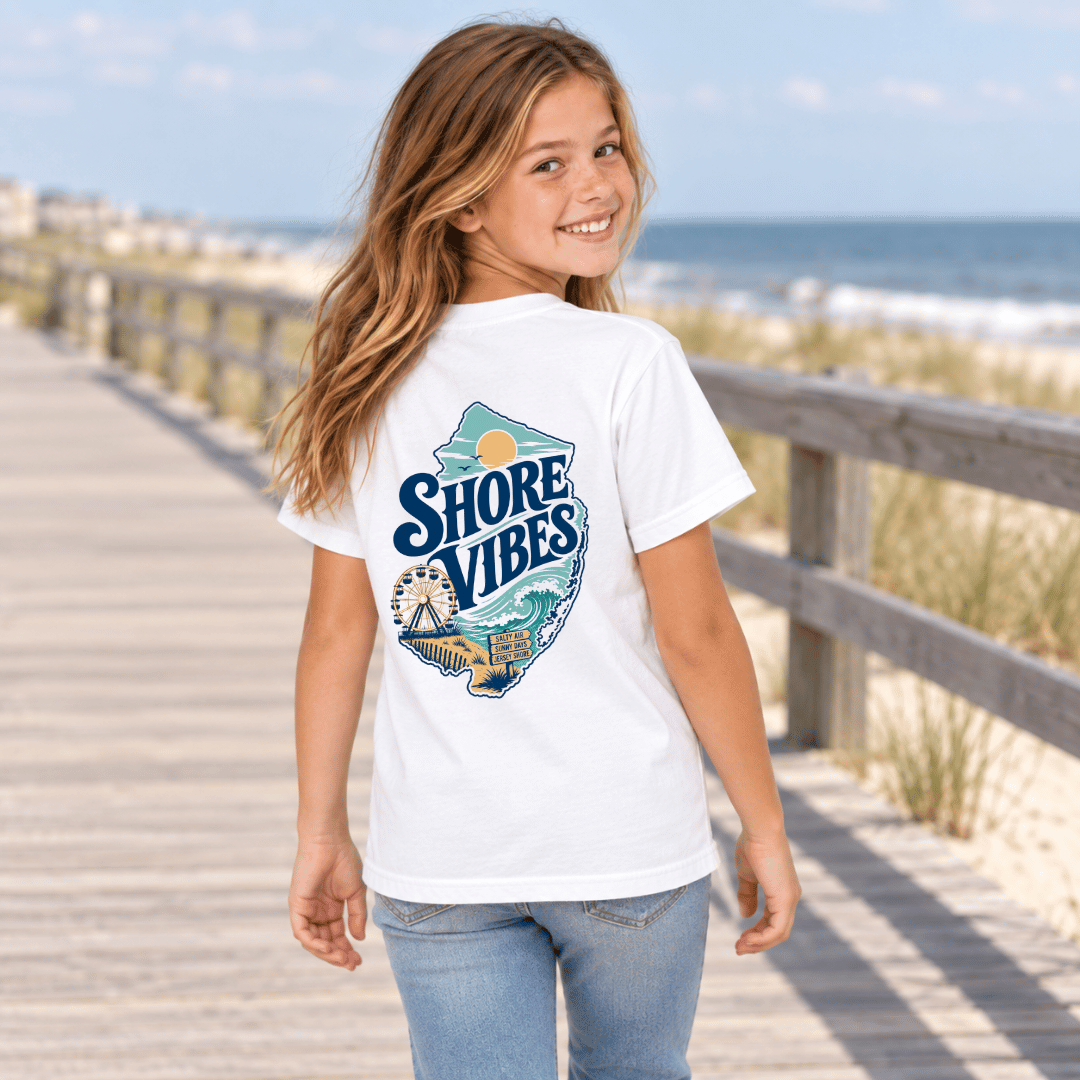 Back view of a girl in a white Shore Vibes t-shirt walking away on a boardwalk path near the beach