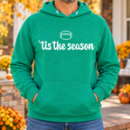 Man wearing a Kelly Green 'tis the season football hoodie on a front porch with fall mums