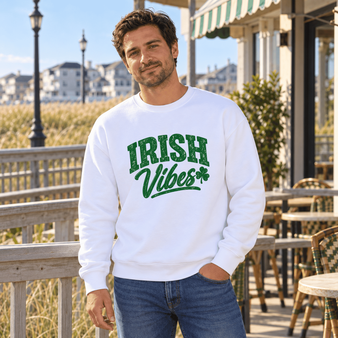 A man wearing a white crewneck sweatshirt with 'IRISH Vibes' printed in green across the chest, standing on an outdoor patio with a seaside background.
