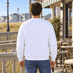 A man viewed from behind wearing a plain white crewneck sweatshirt and blue jeans, standing on a wooden boardwalk with a beach house and ocean in the background.