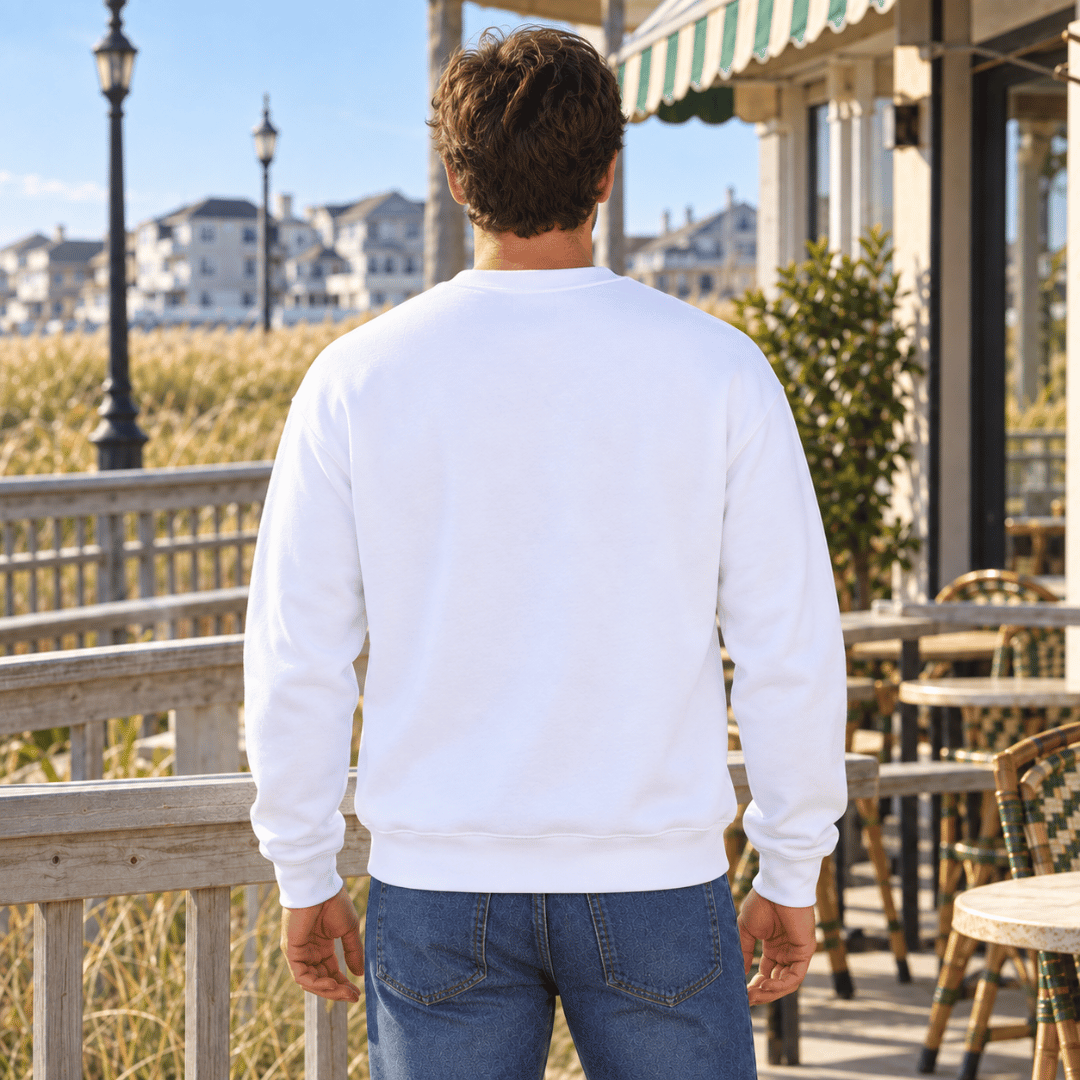 A man viewed from behind wearing a plain white crewneck sweatshirt and blue jeans, standing on a wooden boardwalk with a beach house and ocean in the background.