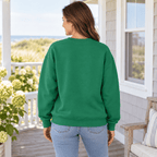 A woman wearing a kelly green crewneck sweatshirt standing, showing the back side of the sweatshirt, on an outdoor patio with tables and a seaside background.
