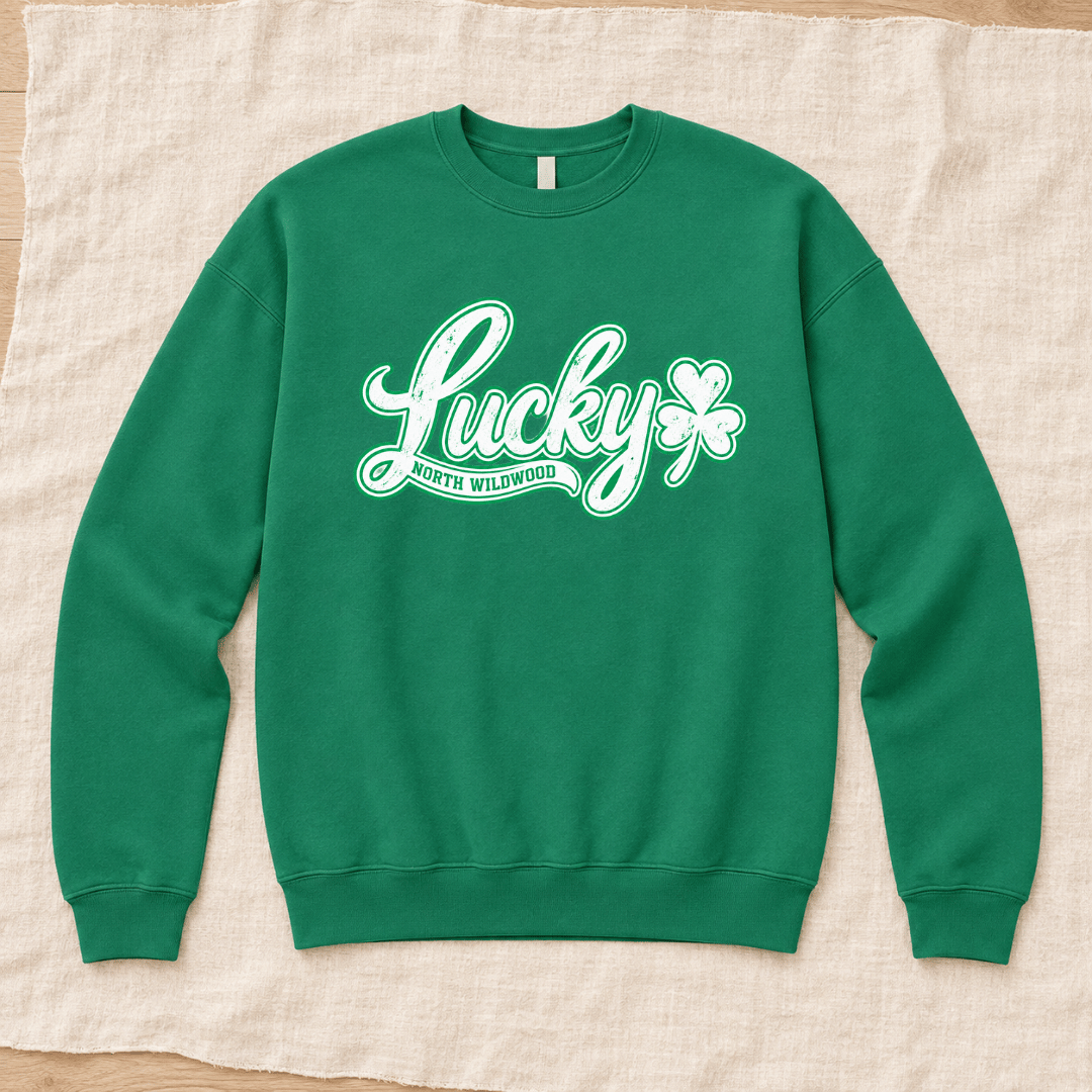 A kelly green crewneck sweatshirt with a white graphic reading 'Lucky North Wildwood' and a shamrock, laid flat on a beige linen fabric background.