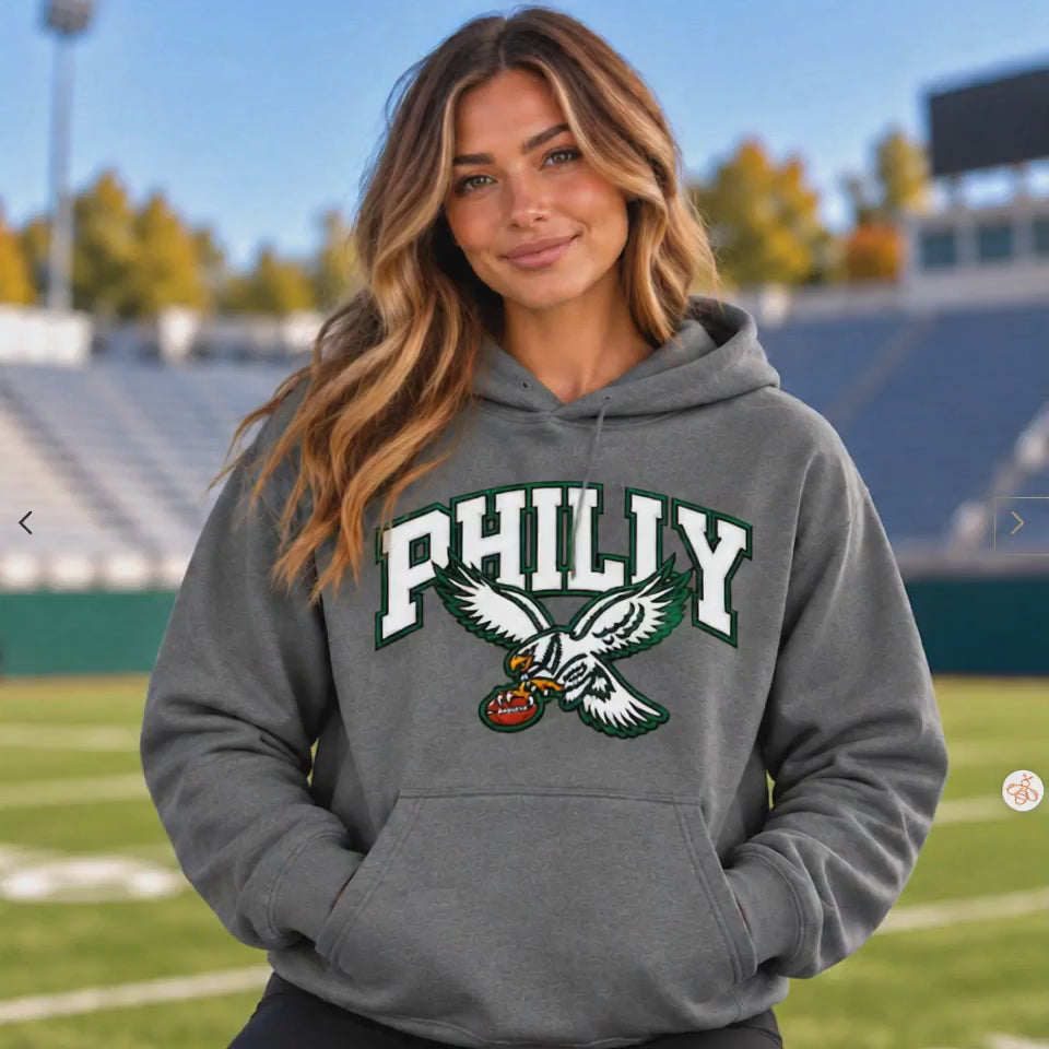 Woman wearing a heather gray hoodie with white and green "PHILLY" varsity lettering and a flying eagle graphic holding a football