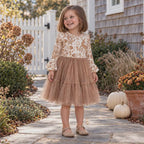 Young girl smiles and gently sways a tan tulle dress with a cream woodland-print bodice and ruffle sleeves beside a coastal cottage decorated with pumpkins and autumn flowers.