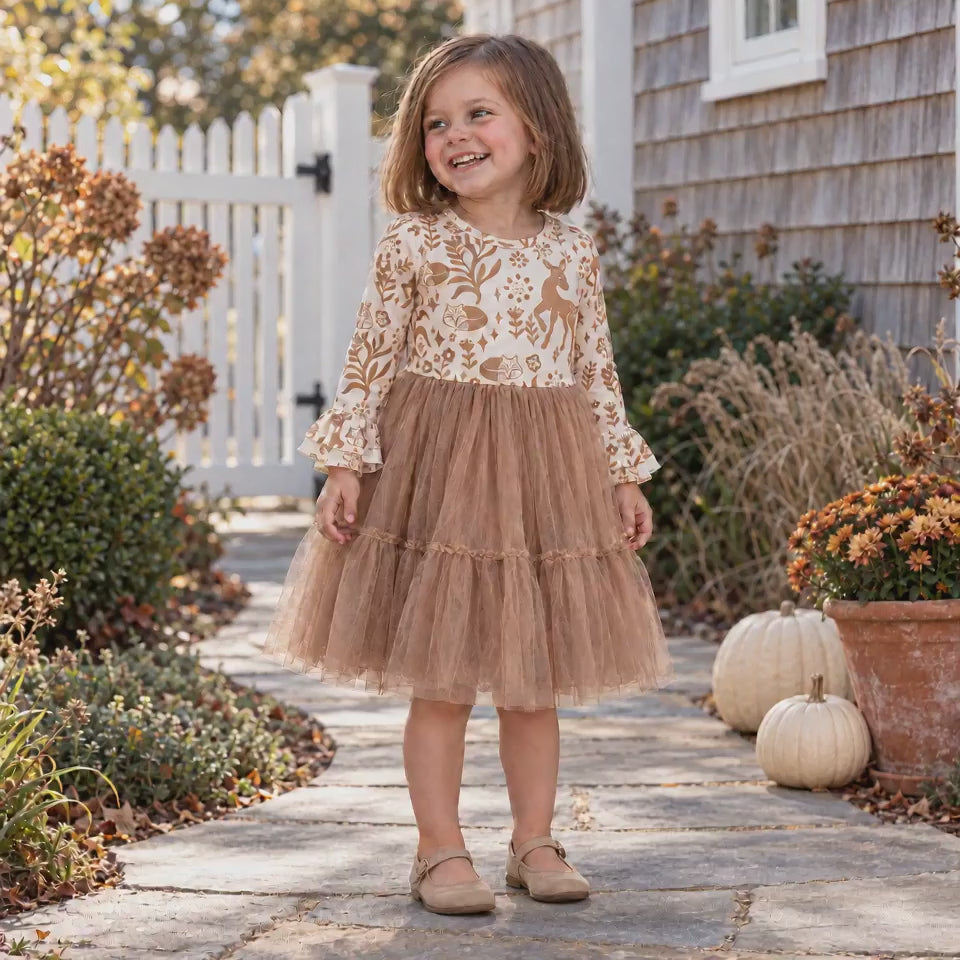 Young girl smiles and gently sways a tan tulle dress with a cream woodland-print bodice and ruffle sleeves beside a coastal cottage decorated with pumpkins and autumn flowers.