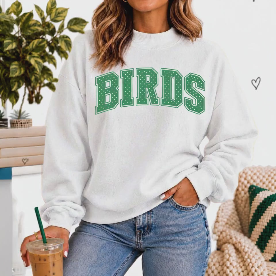 Woman wearing a white mock neck sweatshirt with green polka dot "BIRDS" varsity lettering across the chest