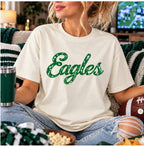 Woman wearing an ivory garment-dyed tee with green distressed "Eagles" script lettering, relaxing on a couch with a tumbler and football on game day