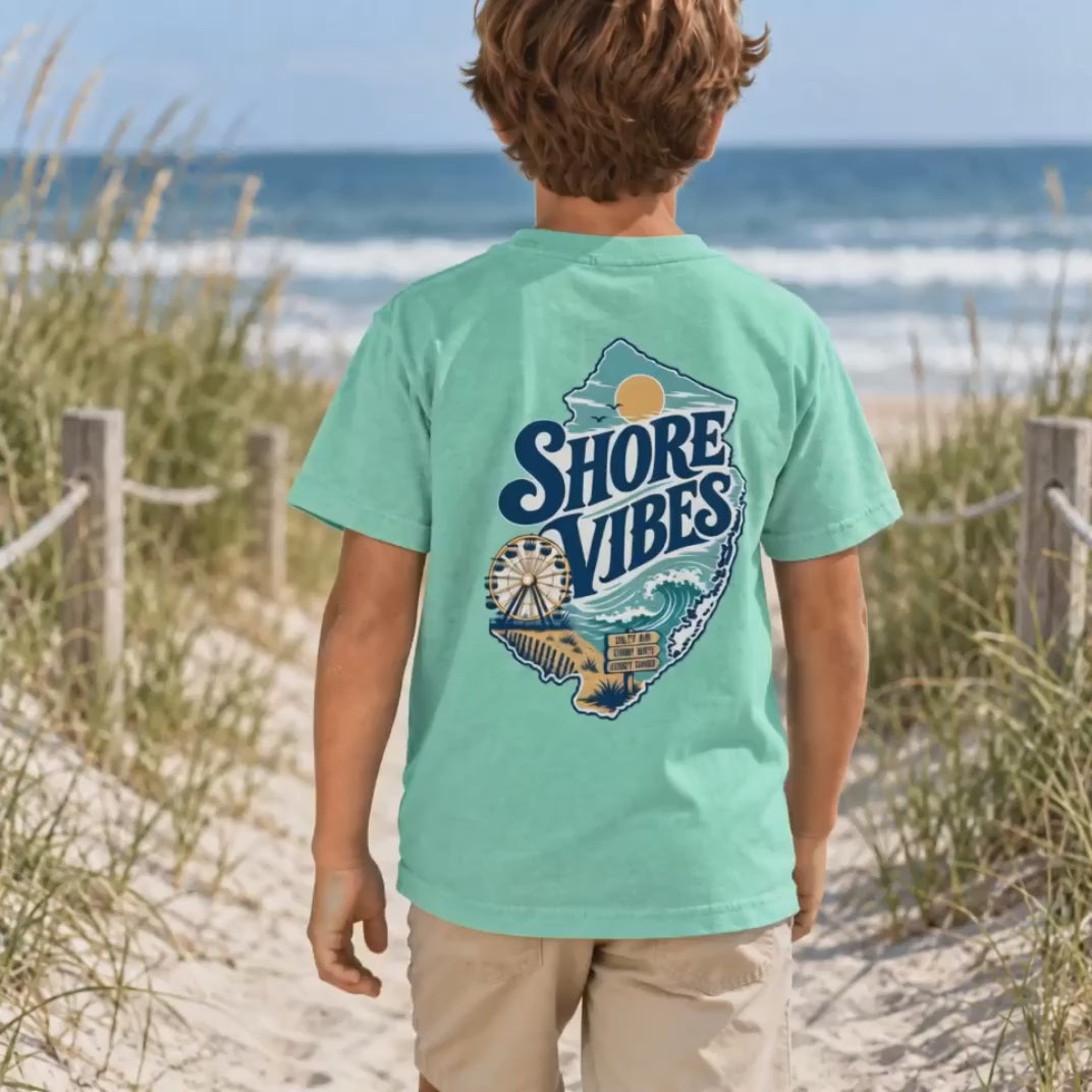 Front and back view of a boy in an Island Reef green Shore Vibes t-shirt standing on a dune path by the ocean