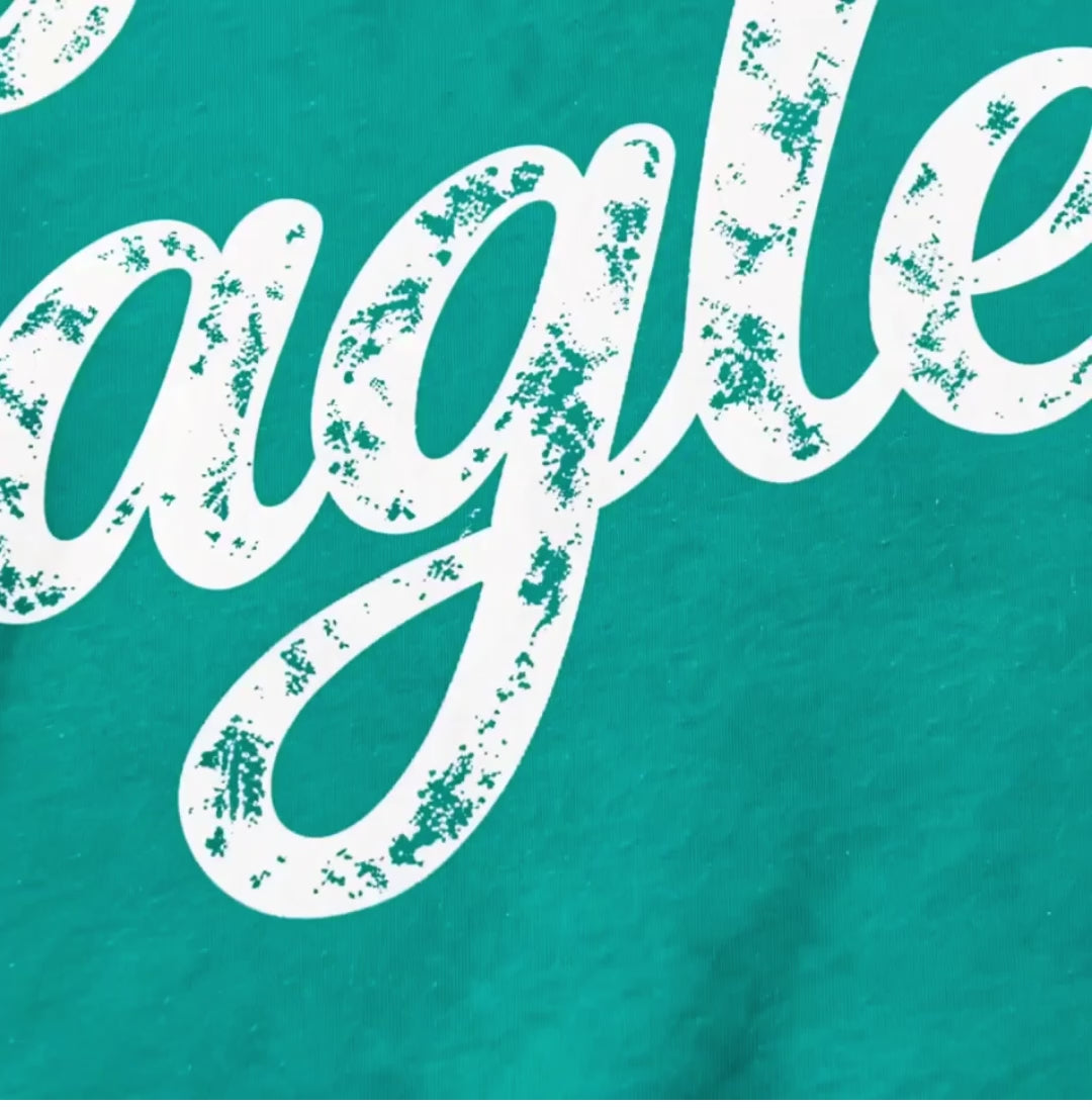 Flat lay of a teal garment-dyed tee with white distressed "Eagles" script lettering across the chest, next to green and white pom-poms